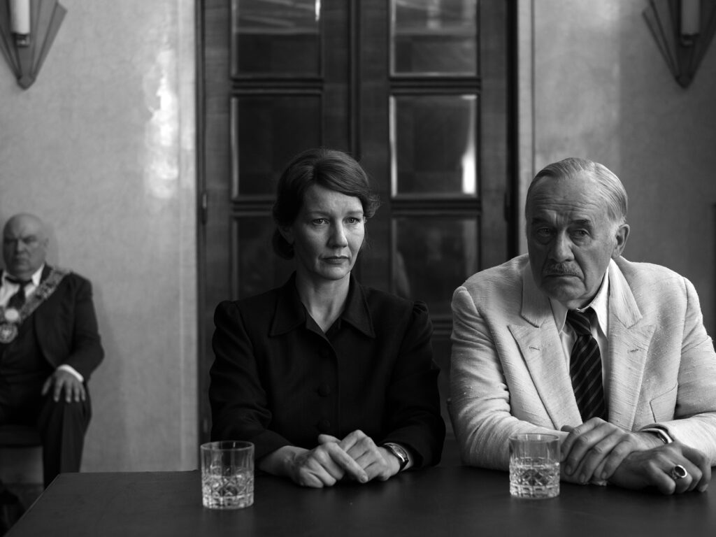 Filmstill aus „Vaterland" von Paweł Pawlikowski – Sandra Hüller und Hanns Zischler sitzen nebeneinander an einem Tisch, im Hintergrund ein Würdenträger mit Amtskette