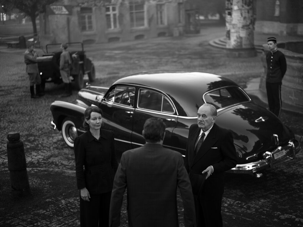 Filmstill aus „Vaterland" von Paweł Pawlikowski – Sandra Hüller und Hanns Zischler neben einem schwarzen Auto, im Hintergrund ein zerstörtes Gebäude