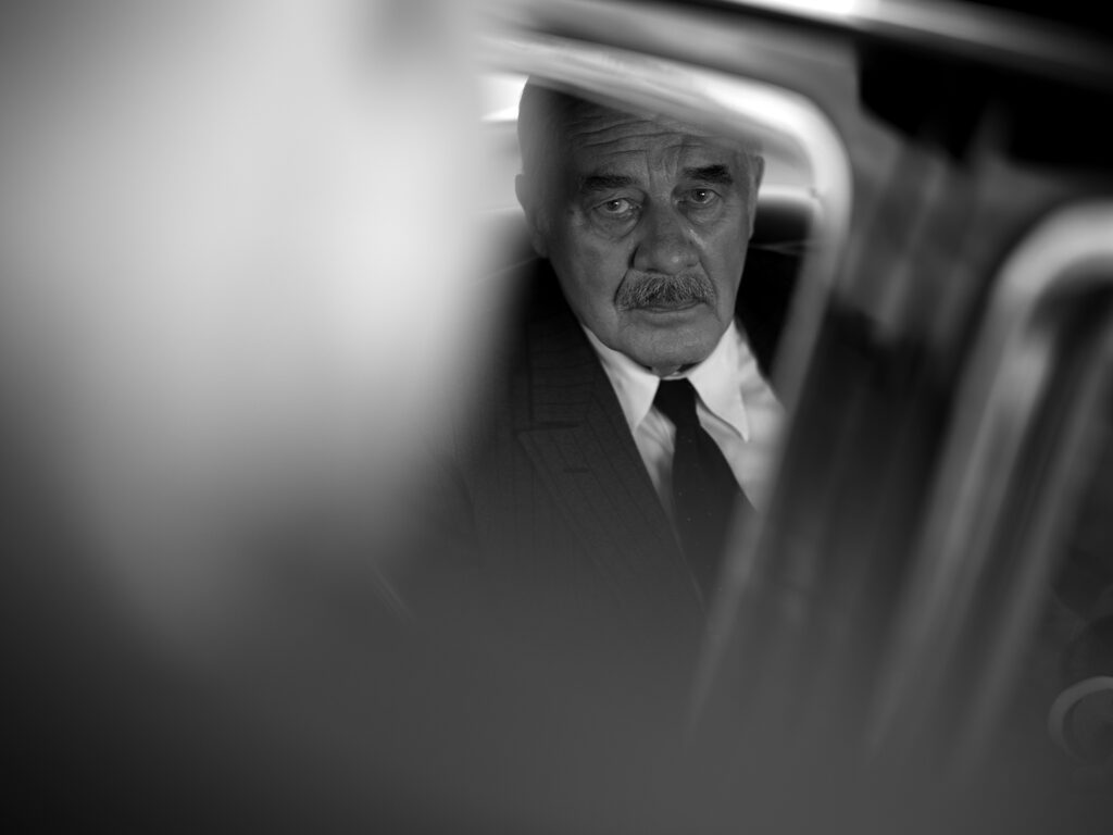 Filmstill aus „Vaterland" von Paweł Pawlikowski – Hanns Zischler als Thomas Mann im Auto, sein Gesicht durch den Fensterrahmen sichtbar, umgeben von unscharfem Vordergrund.