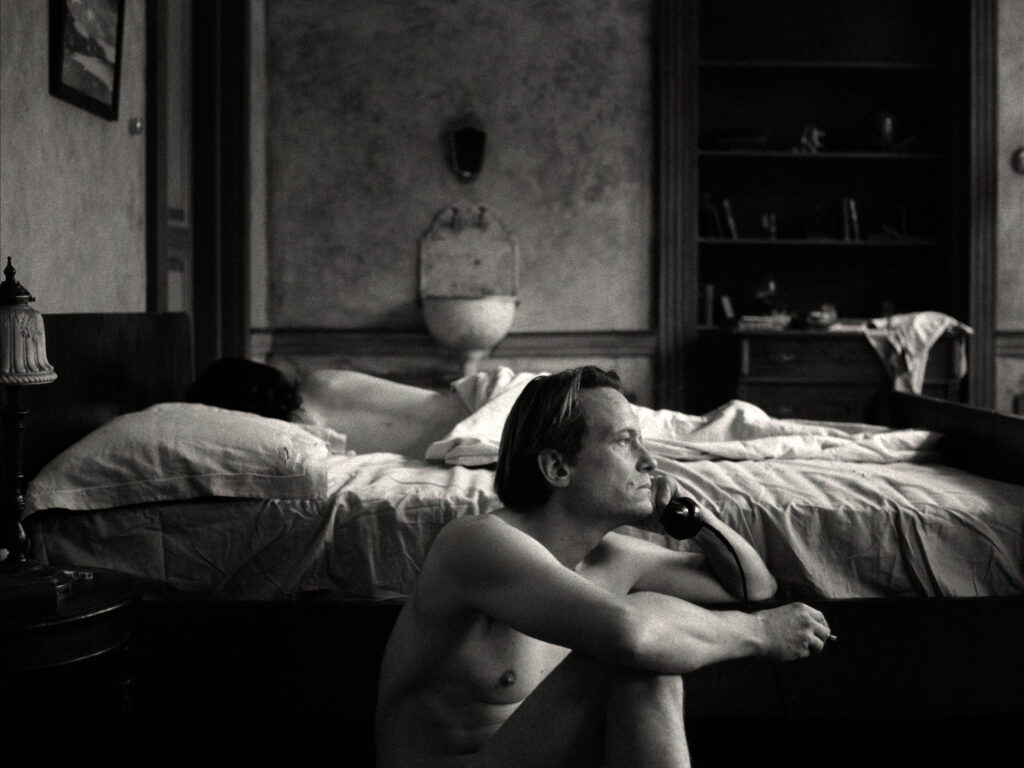 Filmstill aus „Vaterland" – August Diehl als Klaus Mann sitzt mit einer Zigarette am Telefon, im Hintergrund ein Bett mit einer schlafenden Person.
