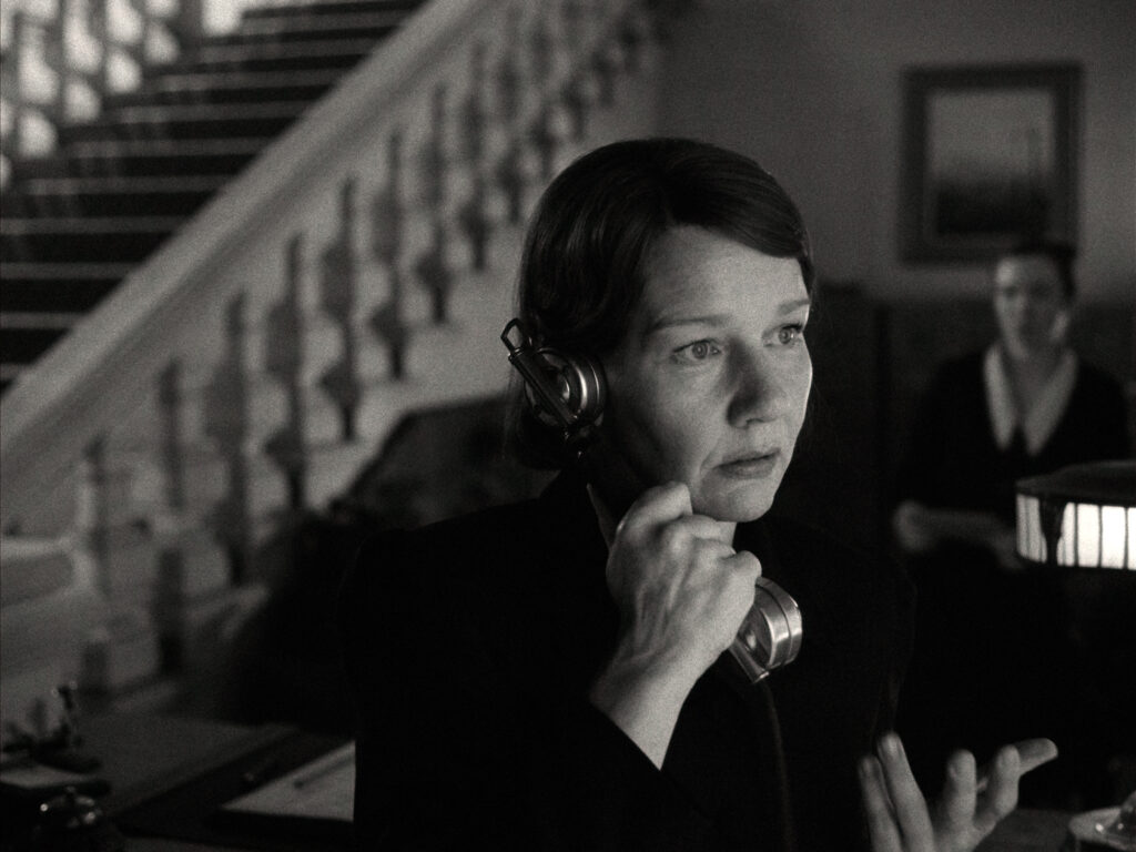 Filmstill aus „Vaterland" – Sandra Hüller als Erika Mann telefoniert, im Hintergrund eine Treppe und eine Person in der Tür.