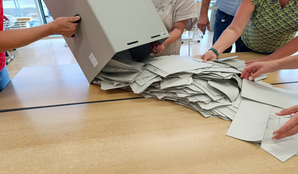 Die Leerung einer Wahlurne, Wahlzettel werden auf einen Tisch ausgeschüttet.