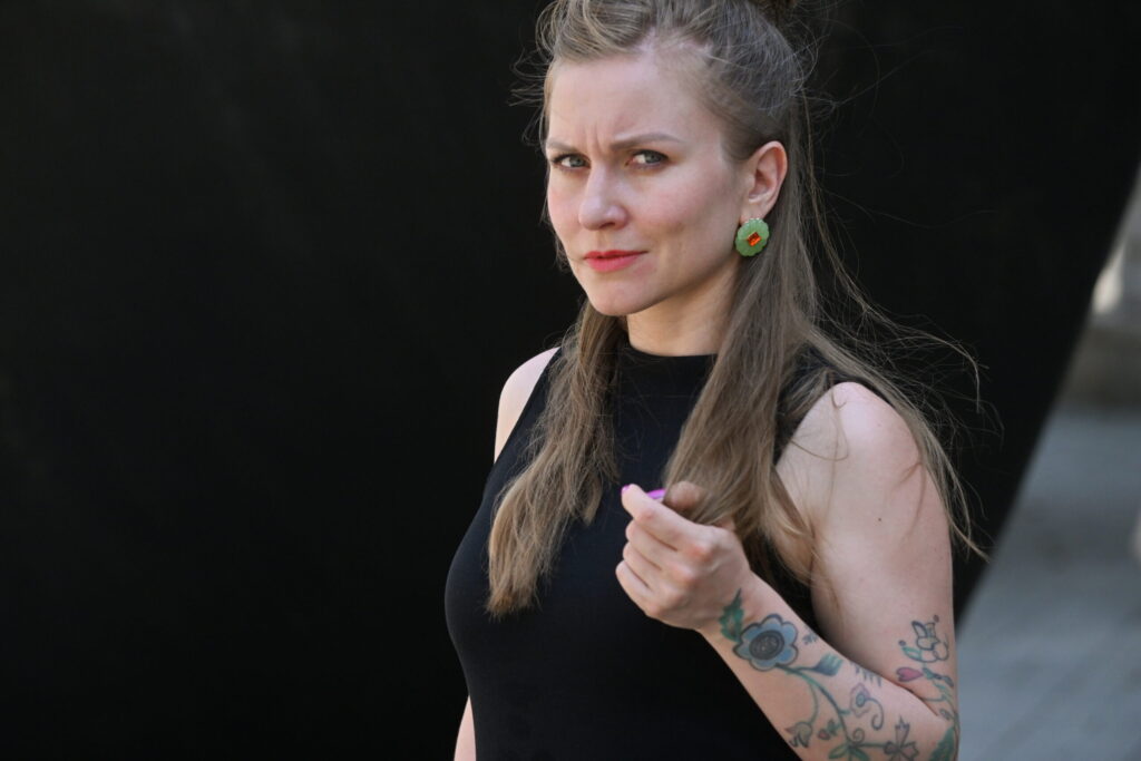 Eine Frau im schwarzen Kleid steht vor einem schwarzen Hintergrund, auf dem linken Arm Tattoos. 