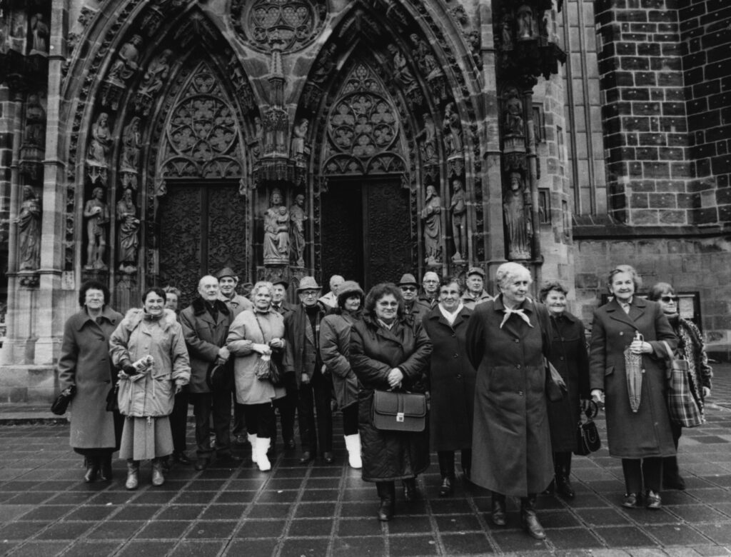 Ein schwarz-weißes Foto zeigt eine Gruppe von älteren Frauen und Männern vor dem Hintergrund eines gewölbten Kircheneingangs. 
