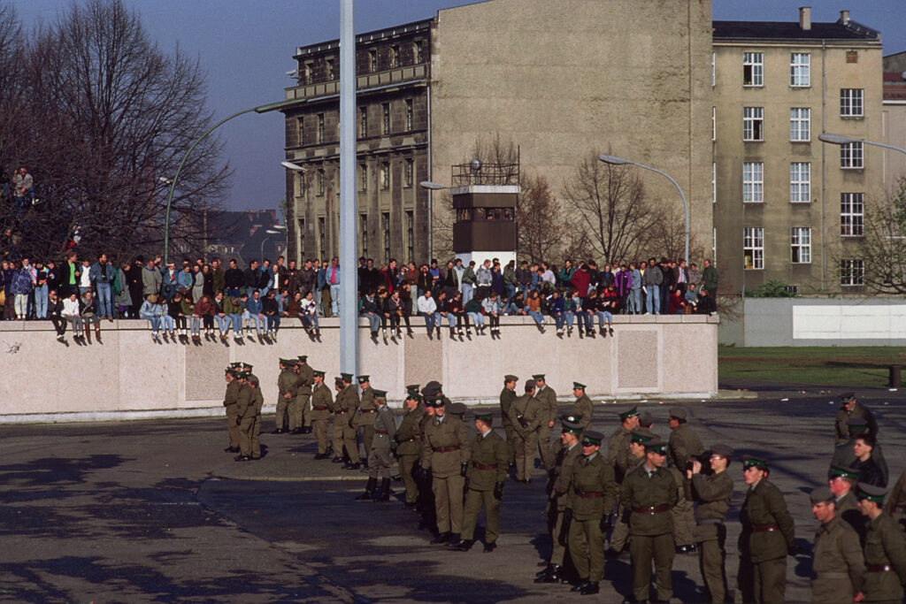 Menschen sitzen auf der Berliner Mauer, im Vordergrund stehen Soldaten.