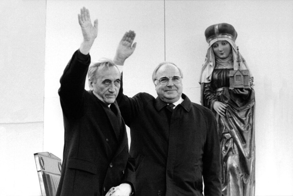 Ein Schwarz-Weiß-Foto zeigt zwei ältere Männer, die winken. Rechts von ihnen eine Heilige-Maria-Figur.