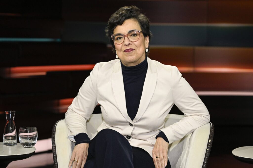 Eine schwarz-weiß gekleidete Frau mit Brille sitzt in einem Fernsehstudio und schaut in die Kamera. 