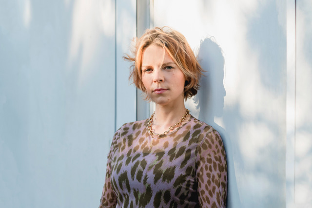 Eine Frau, angelehnt an eine Wand, schaut in die Kamera.