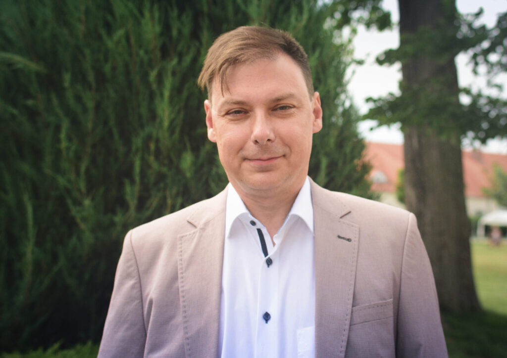Ein Mann im hellen Jackett steht im Freien, im Hintergrund grüne Bäume.