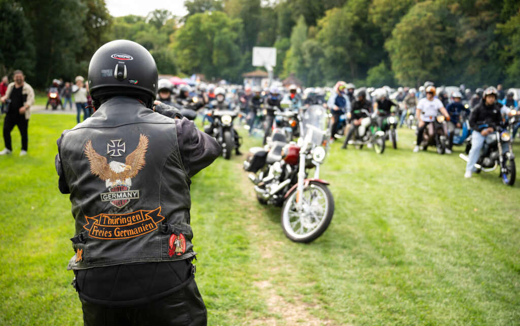 Der Rücken eines Mannes in einer Lederjacke mit der Aufschrift Germania Thüringen, im Hintergrund mehrere Mopeds der Marke Simson.