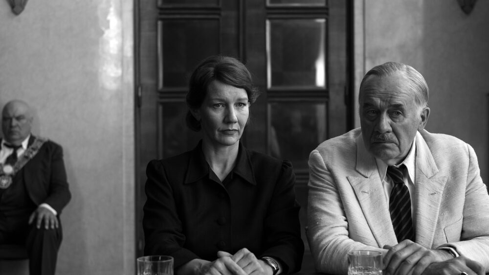 Filmstill aus „Vaterland" von Paweł Pawlikowski – Sandra Hüller und Hanns Zischler sitzen nebeneinander an einem Tisch, im Hintergrund ein Würdenträger mit Amtskette