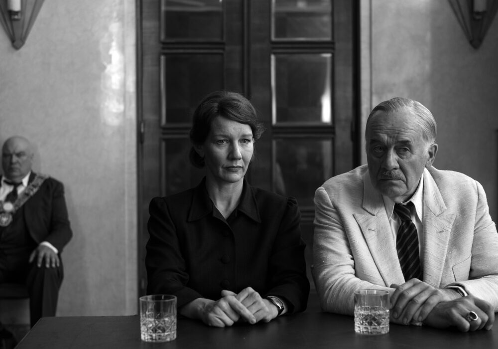 Filmstill aus „Vaterland" von Paweł Pawlikowski – Sandra Hüller und Hanns Zischler sitzen nebeneinander an einem Tisch, im Hintergrund ein Würdenträger mit Amtskette