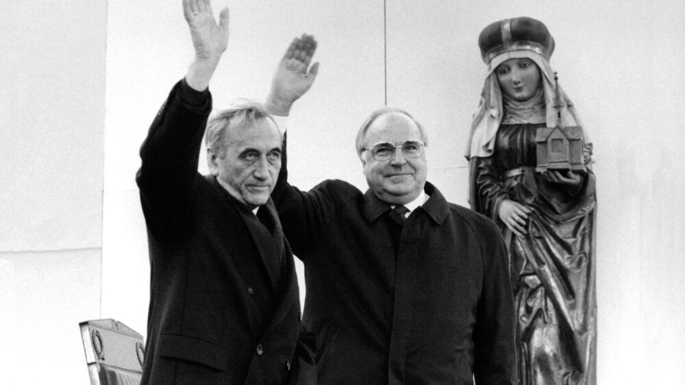 Ein Schwarz-Weiß-Foto zeigt zwei ältere Männer, die winken. Rechts von ihnen eine Heilige-Maria-Figur.