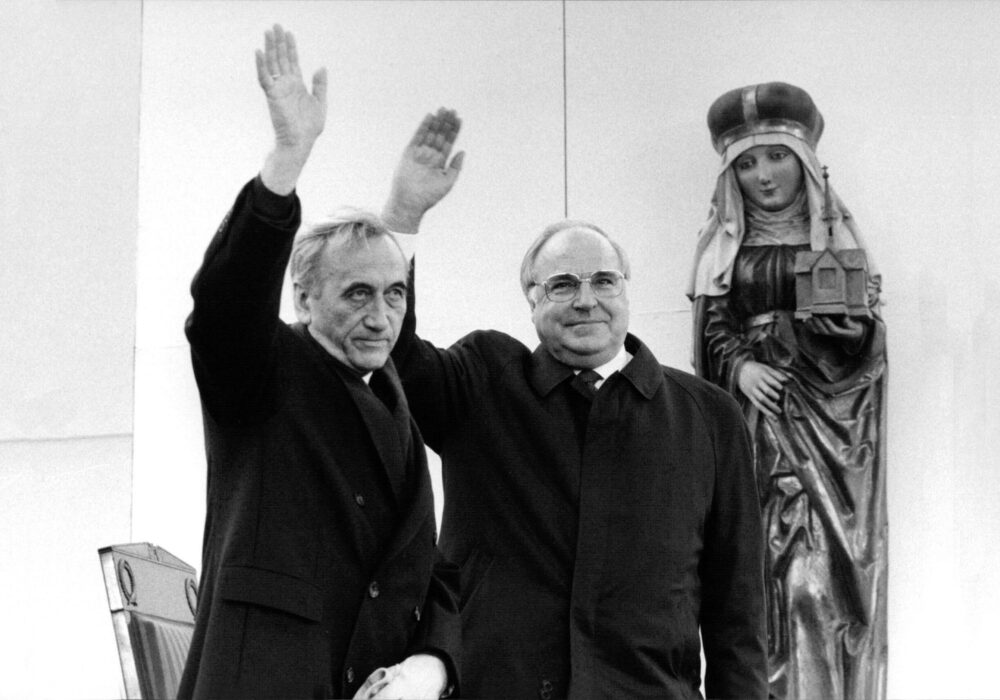 Ein Schwarz-Weiß-Foto zeigt zwei ältere Männer, die winken. Rechts von ihnen eine Heilige-Maria-Figur.