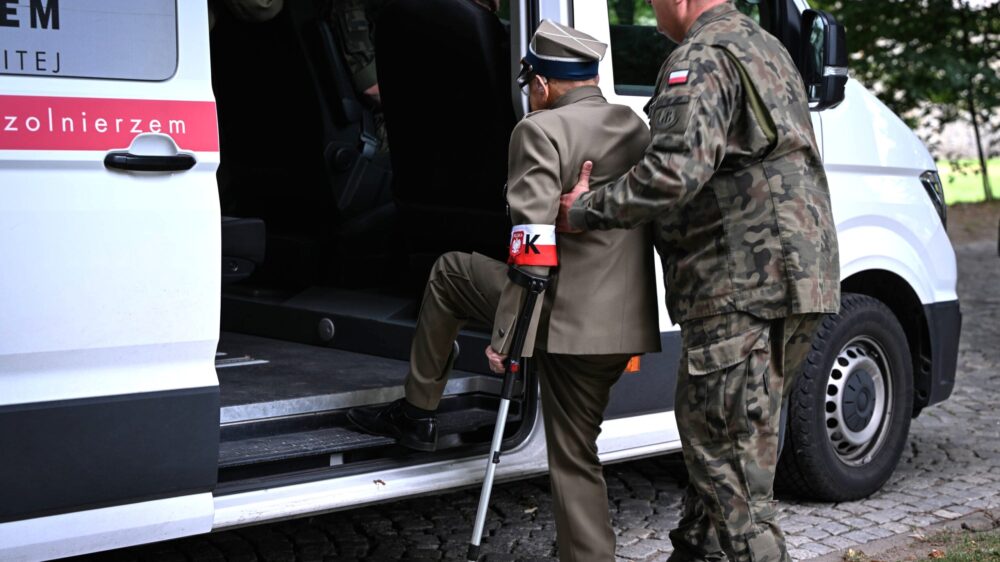 Ein hochbetagter Mann in Uniform und mit Gehstock steigt in ein Auto, ein Soldat hilft ihm dabei.