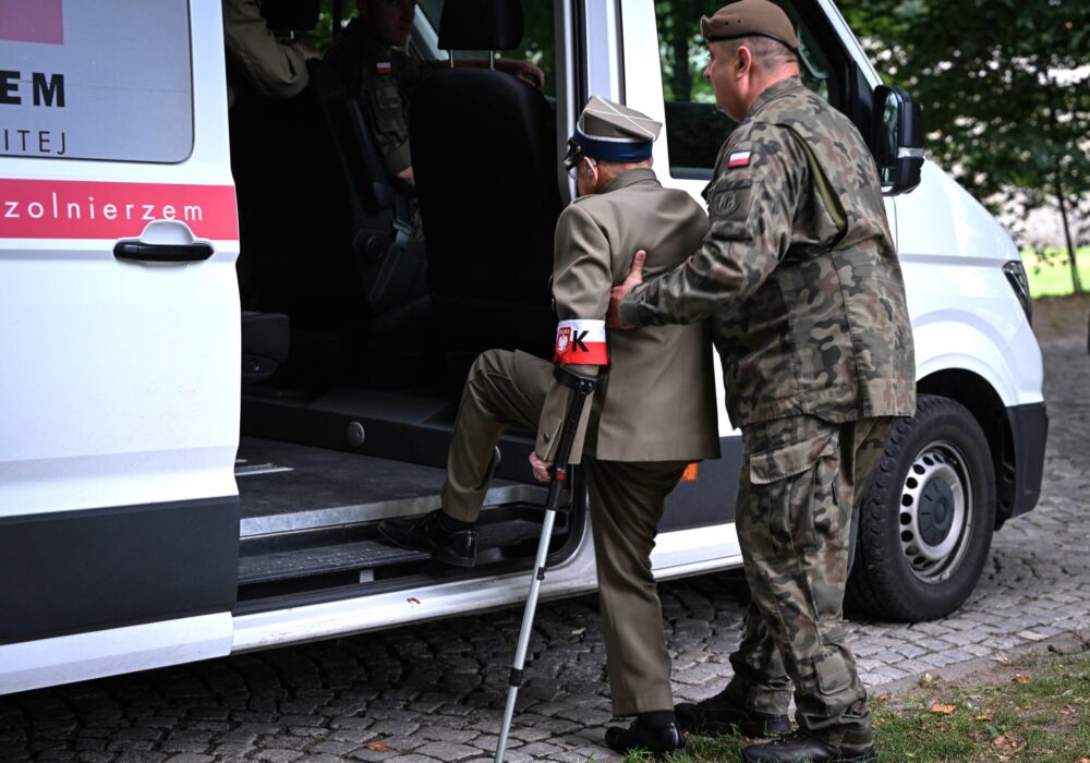 Ein hochbetagter Mann in Uniform und mit Gehstock steigt in ein Auto, ein Soldat hilft ihm dabei.